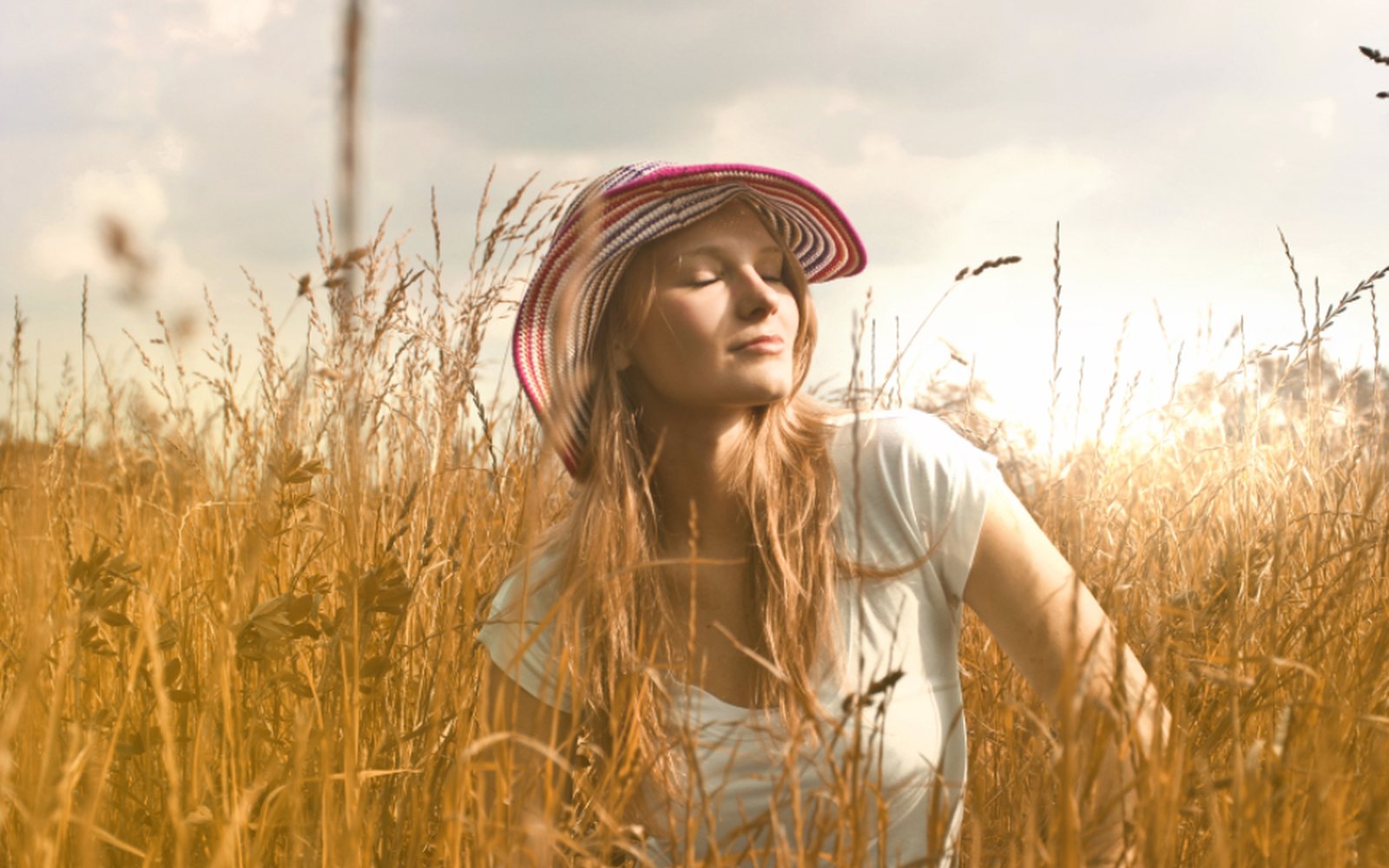 A woman in a sun hat with her eyes closed, standing in tall golden grass in warm sunlight